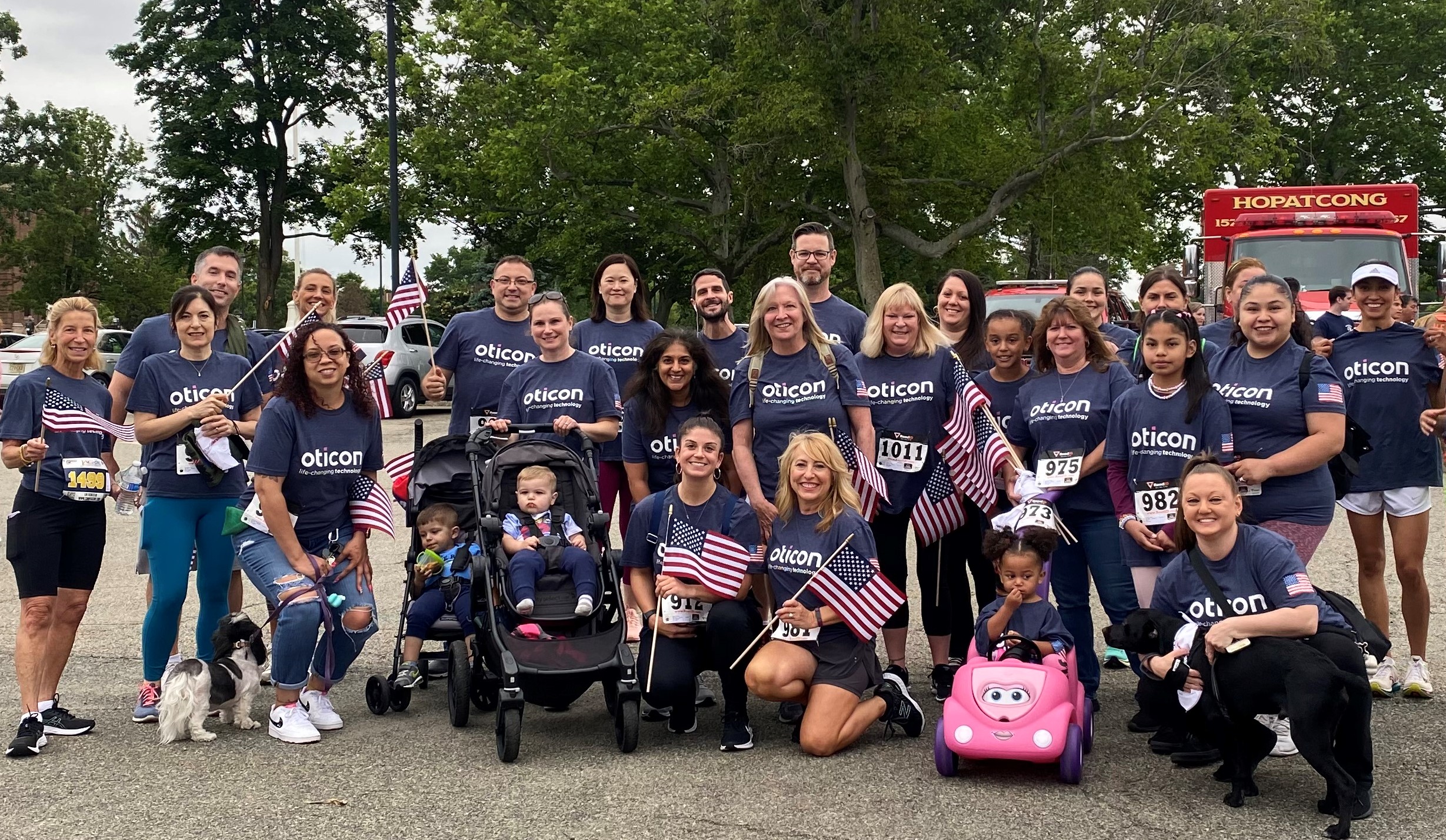 Group photo of people wearing Oticon shirts and holding small flags in park like setting.