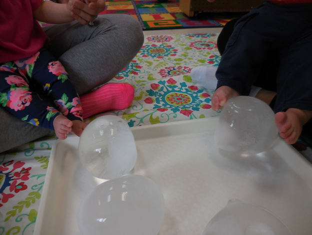 Two small children seated on the floor playing with large ice balls on a plastic tray 