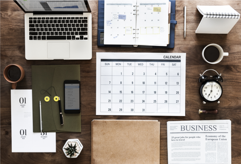 Overhead view of desktop with laptop calendars newspaper coffee pens phone and alarm clock