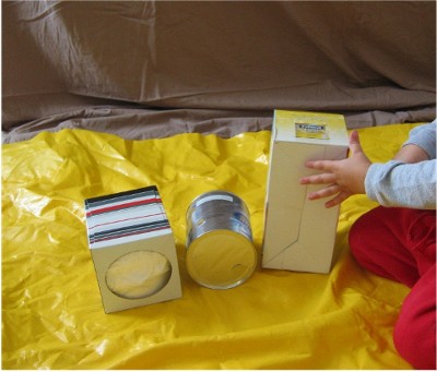 Child seated on yellow tarp playing with two recycled boxes and one can. 